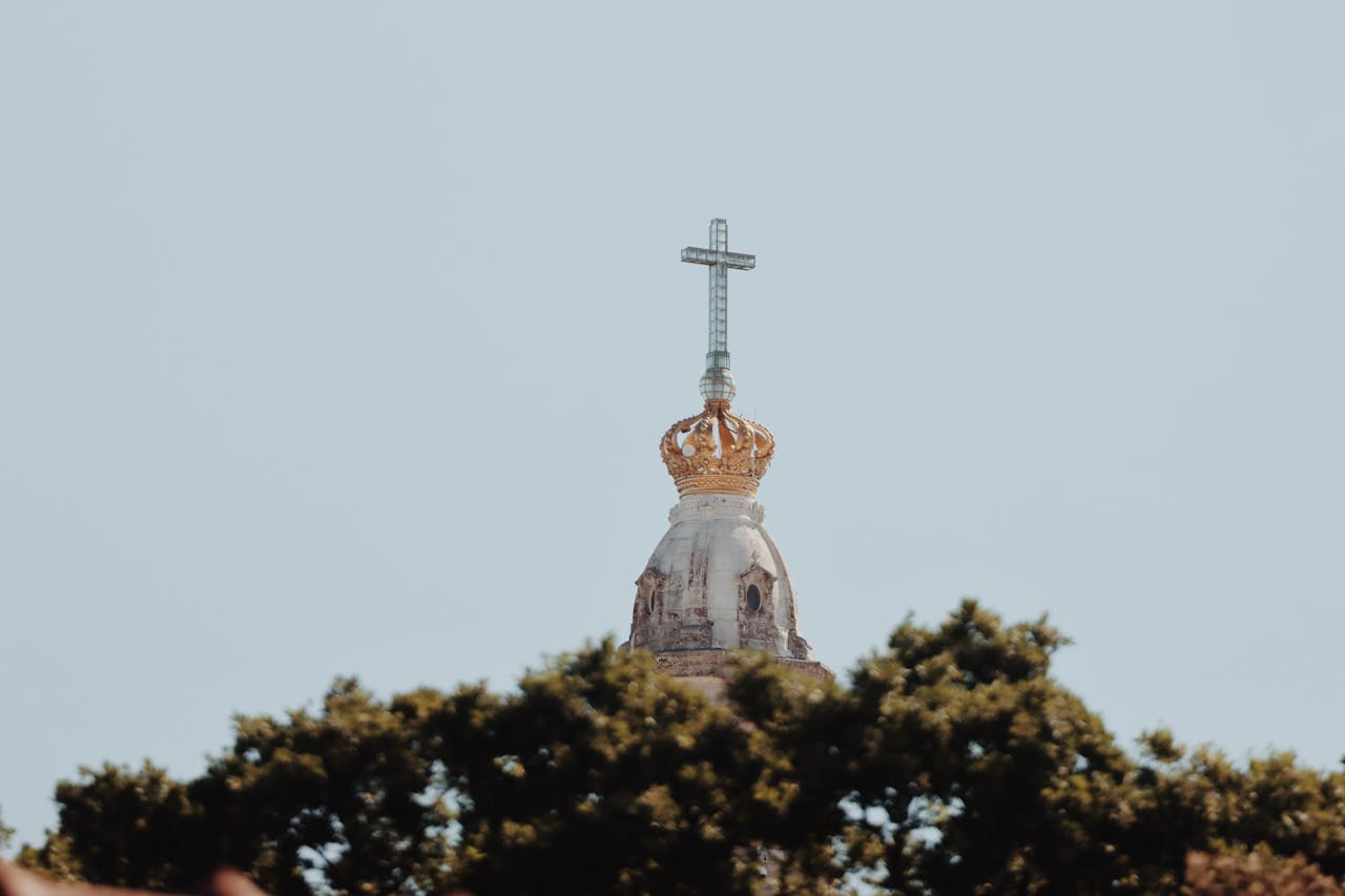 A serene view of the cross atop the sanctuary tower in Fátima, Portugal, surrounded by greenery.